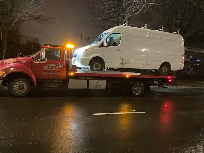 Flatbed carrying a white Sprinter cargo van on a rain-slicked Chicago street at night